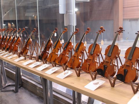 A row of violins on stands along a workbench, each with a maker's card beside it